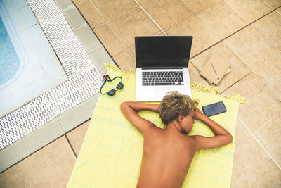 High angle view of boy sleeping by laptop and phone on beach towel at poolside