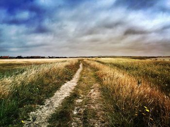 Dirt road passing through field against cloudy sky