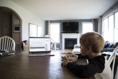 Wide side view of blonde boy eating cereal watching lesson on laptop