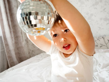 Little boy plays with sparkling mirror disco ball in bed and laughing happily. joyful toddler. 