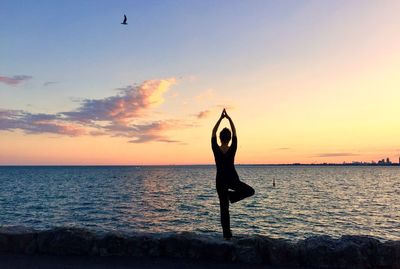 Rear view of woman practicing tree pose yoga on retaining wall against sea during sunset