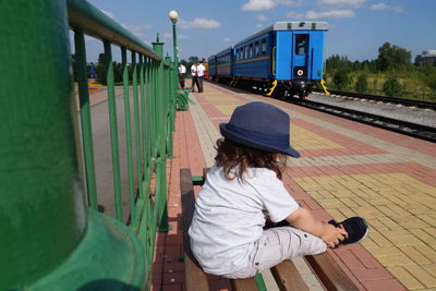 Rear view of girl sitting on bench at railroad station against sky