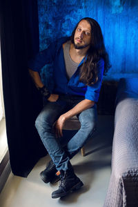 Man with long hair and beard sits at home on a chair by the window in a blue shirt and jeans