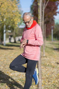 Senior man in sports clothing checking time on smart watch while leaning on tree