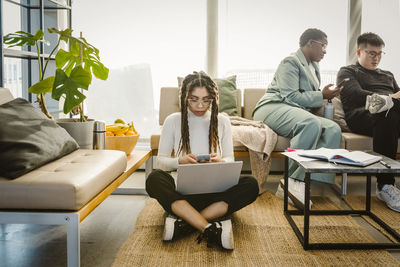 Programmer using mobile phone while sitting with laptop on carpet in creative office