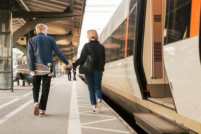 Rear view of young couple holding hands while walking by train at railroad station platform