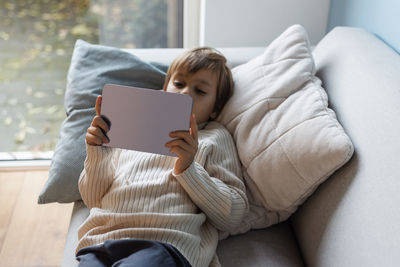 Little cute boy watching cartoons and playing games on a digital tablet lying on the sofa.