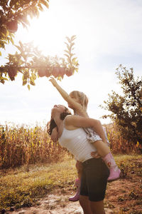 Girl piggybacking sister for reaching apples on trees in orchard