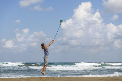 Full length of girl jumping with fishing net at beach against sky