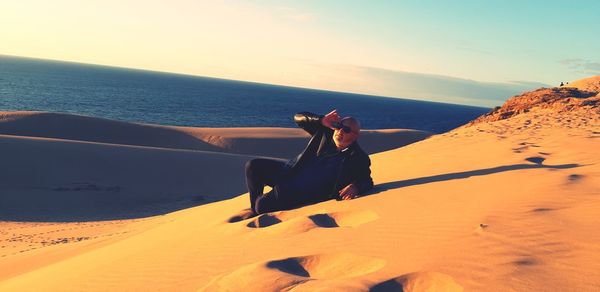 Mature man with sunglasses lying on sand at beach against sky during sunset