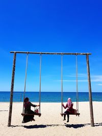 Rear view of women swinging at beach against sky on sunny day