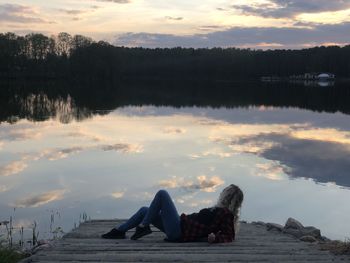 Man lying down on lake against sky during sunset