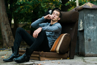 Young man smoking while sitting on sofa against trees