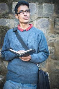 Thoughtful man holding book while standing against stone wall