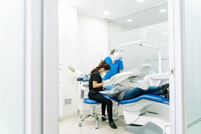 Concentrated female stomatologist in mask sitting near dental chair while treating client with help of nurse in light cabinet