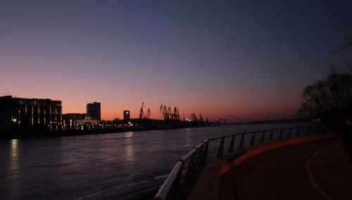 Silhouette buildings by river against clear sky during sunset