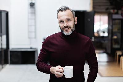 Portrait of smiling mature businessman having coffee while standing at creative office