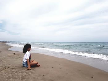 Side view of young woman sitting at seashore against sky