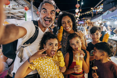 Cheerful man taking selfie with family enjoying street food at market on vacation
