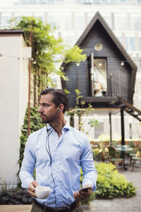 Thoughtful businessman holding coffee cup and smart phone at office yard