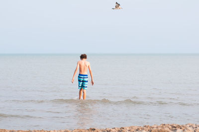 Rear view of shirtless boy standing in sea against clear sky