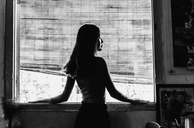 Rear view of woman standing against window at home