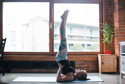 Full length of woman performing shoulder stand by window at home