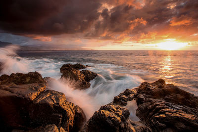 Scenic view of sea waves splashing against rock formation during sunset