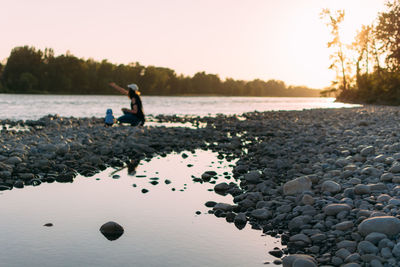 Mother and kid crouching against lake during sunset