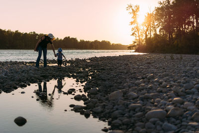 Mother and son walking at riverbank against clear sky during sunset