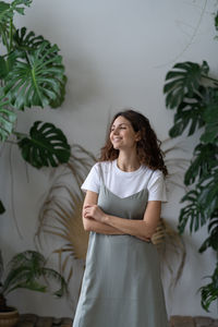 Young happy female gardener standing against lush monstera deliciosa houseplants in home garden