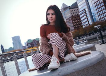 Young woman sitting on seat against buildings in city