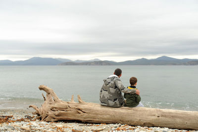Father and son sitting on an old tree without bark on a sandy beach and admiring the blue clear sea 