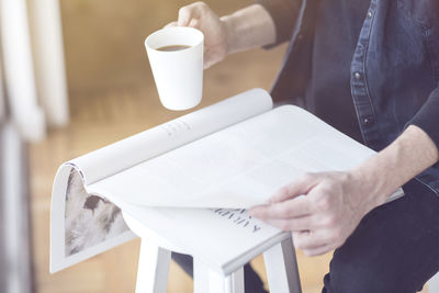 Midsection of man holding coffee reading magazine while sitting at home