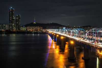 Illuminated bridge over river against buildings at night