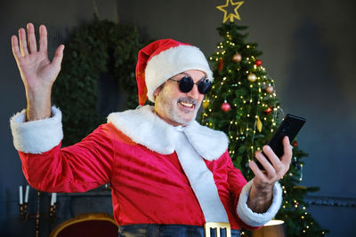 Close-up of smiling man wearing santa costume using mobile phone against christmas tree