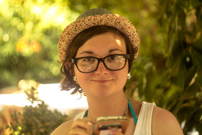 Close-up portrait of woman in eyeglasses holding mobile phone against tree