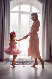 Mom and daughter baby in pink dresses sitting on the floor of the house, background of the window