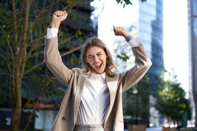 Portrait of young woman with arms raised standing against trees