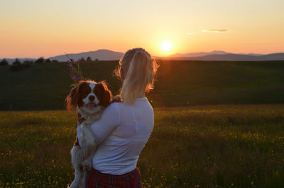 Woman holding her cavalier king charles spaniel dog and looking at hills in landscape in sunset