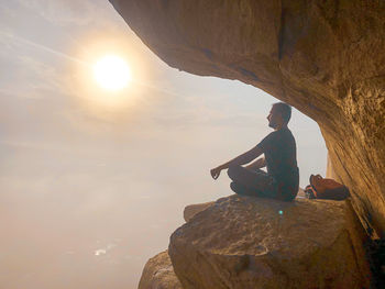 Man sitting on rock against sky during sunset