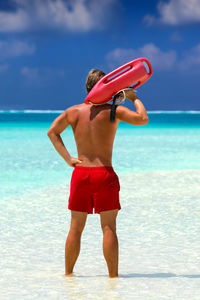 Rear view of lifeguard holding rescue can with hand on hip standing at beach against sky