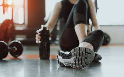 Low section of woman holding water bottle while sitting on floor in gym