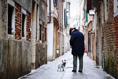 Rear view of man walking dog in alley between buildings in city