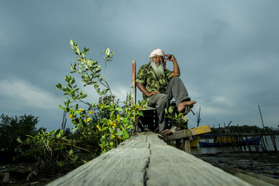 Low angle portrait of senior man sitting on wood against cloudy sky