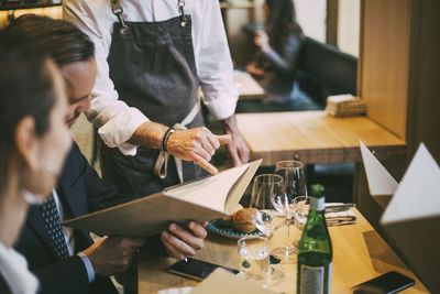 Midsection of waiter pointing at menu while business professionals sitting in restaurant