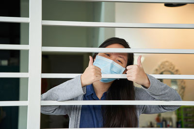 Portrait of young woman with face mask with thums up behind window