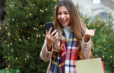 Smiling woman using mobile phone while standing against christmas tree