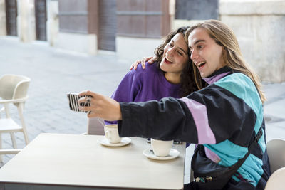 Friends taking selfies with a mobile phone while sitting at an outdoor cafe.