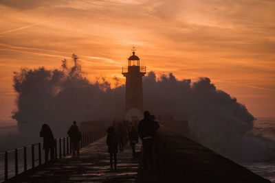People by lighthouse amidst sea against sky during sunset